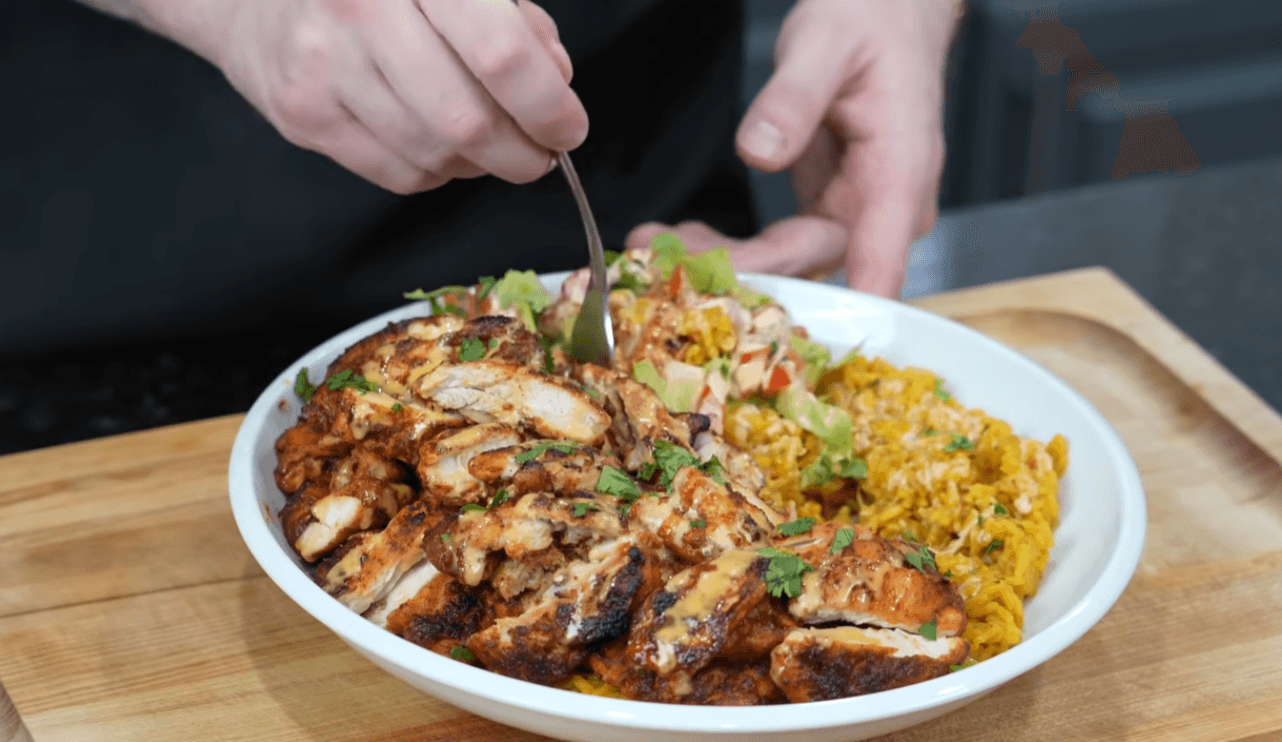A close-up photograph of a white bowl filled with sliced grilled chicken breast topped with a creamy sauce. The dish is served with seasoned yellow rice and a fresh cucumber, tomato, and cilantro salad. The bowl rests on a wooden cutting board against a dark background.