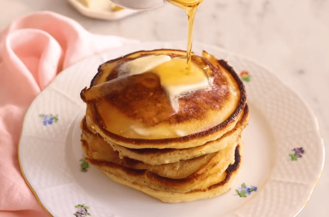 A tall stack of freshly cooked pancakes from our fluffy homemade pancakes recipe, topped with a pat of melting butter and dripping with maple syrup.